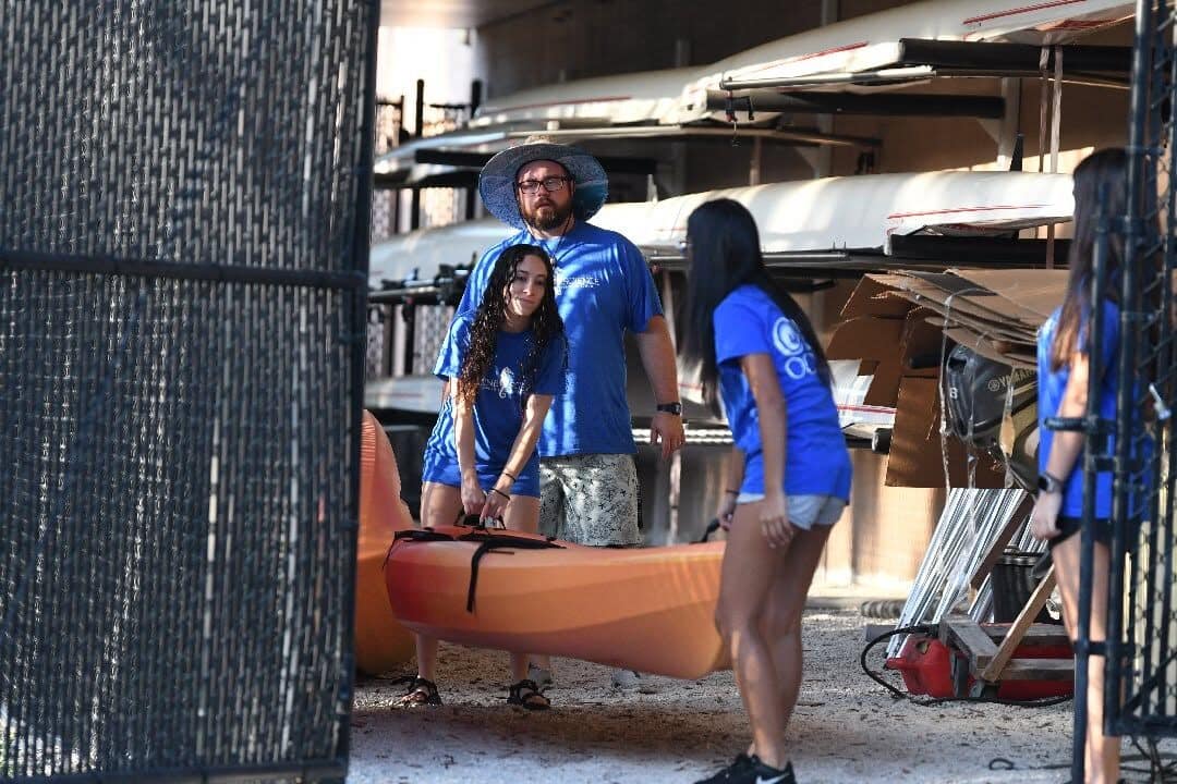 Students and an adult moving a kayak beside storage racks.