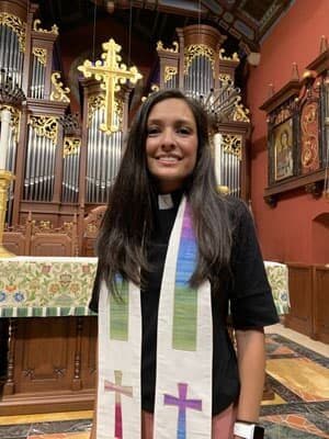 The school chaplain, wearing a clergy stole, stands in the chapel before the organ pipes and altar.