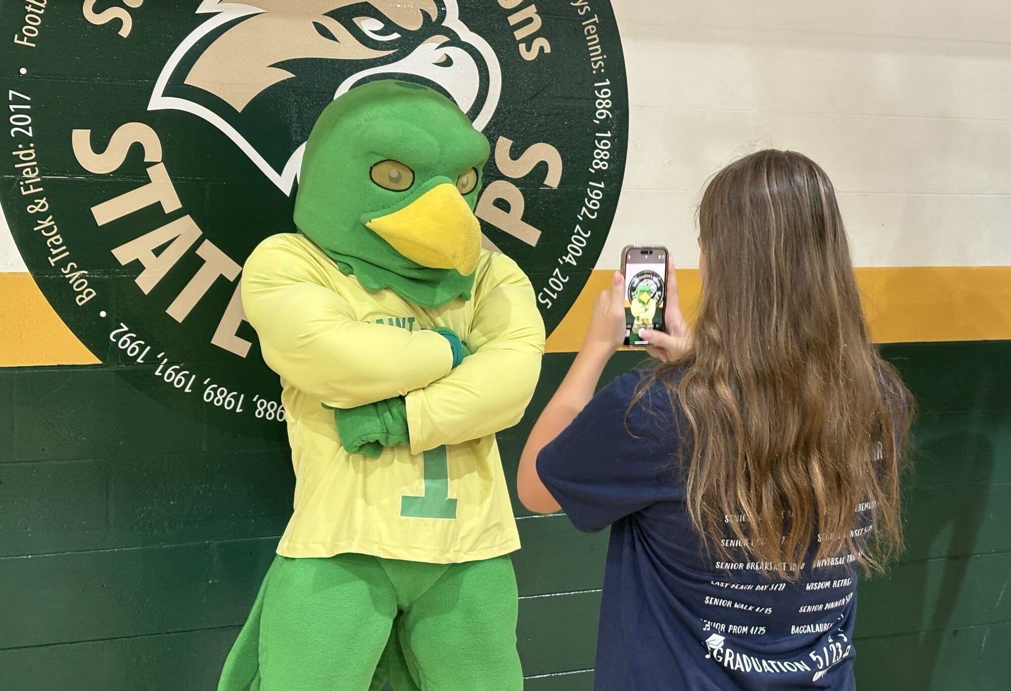 A student photographs the Saint Stephen's Falcon mascot in the gymnasium in front of the athletics championship banner.