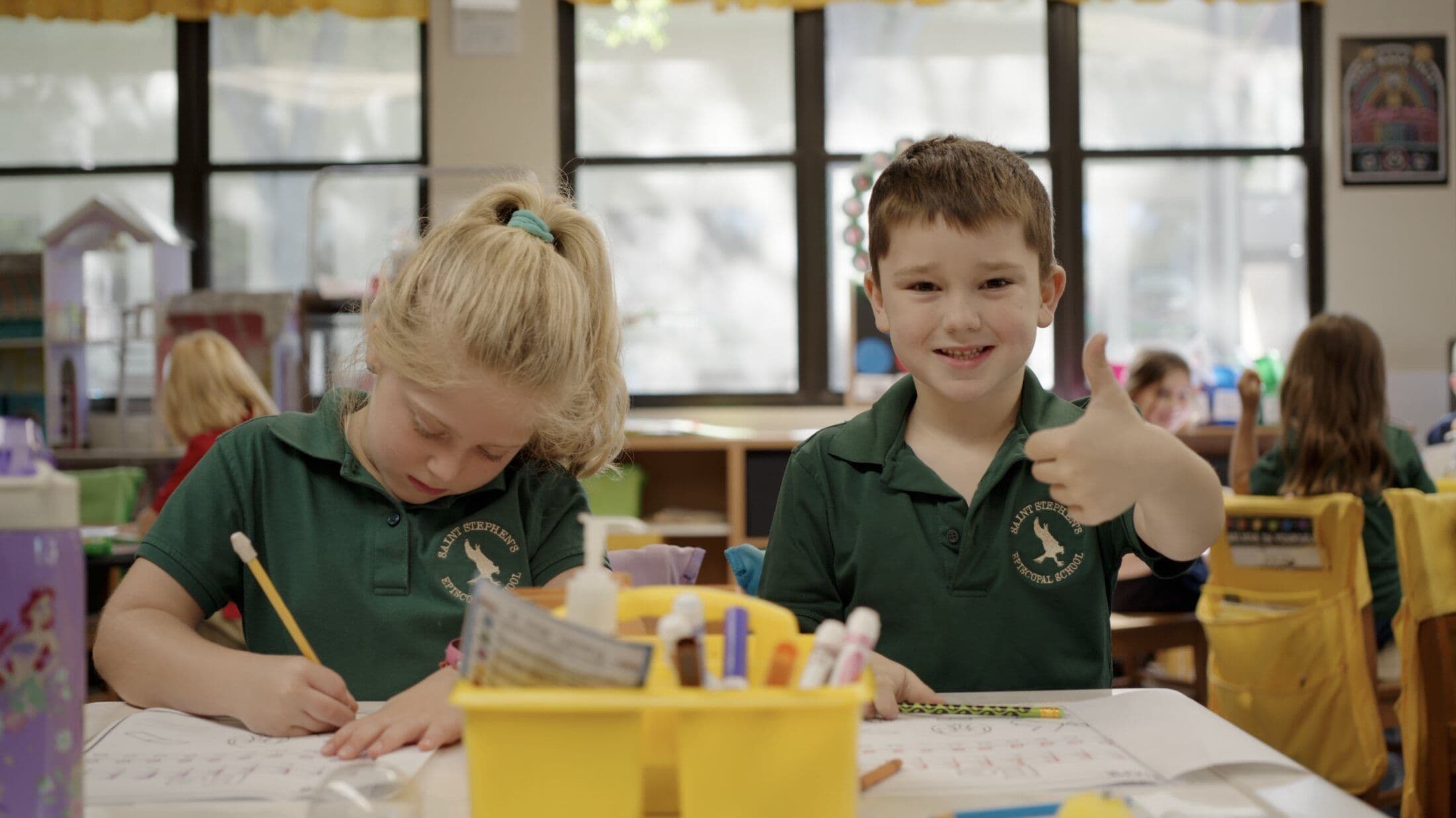 Two Lower School students in green polos work on writing exercises at a table, one giving a thumbs-up.