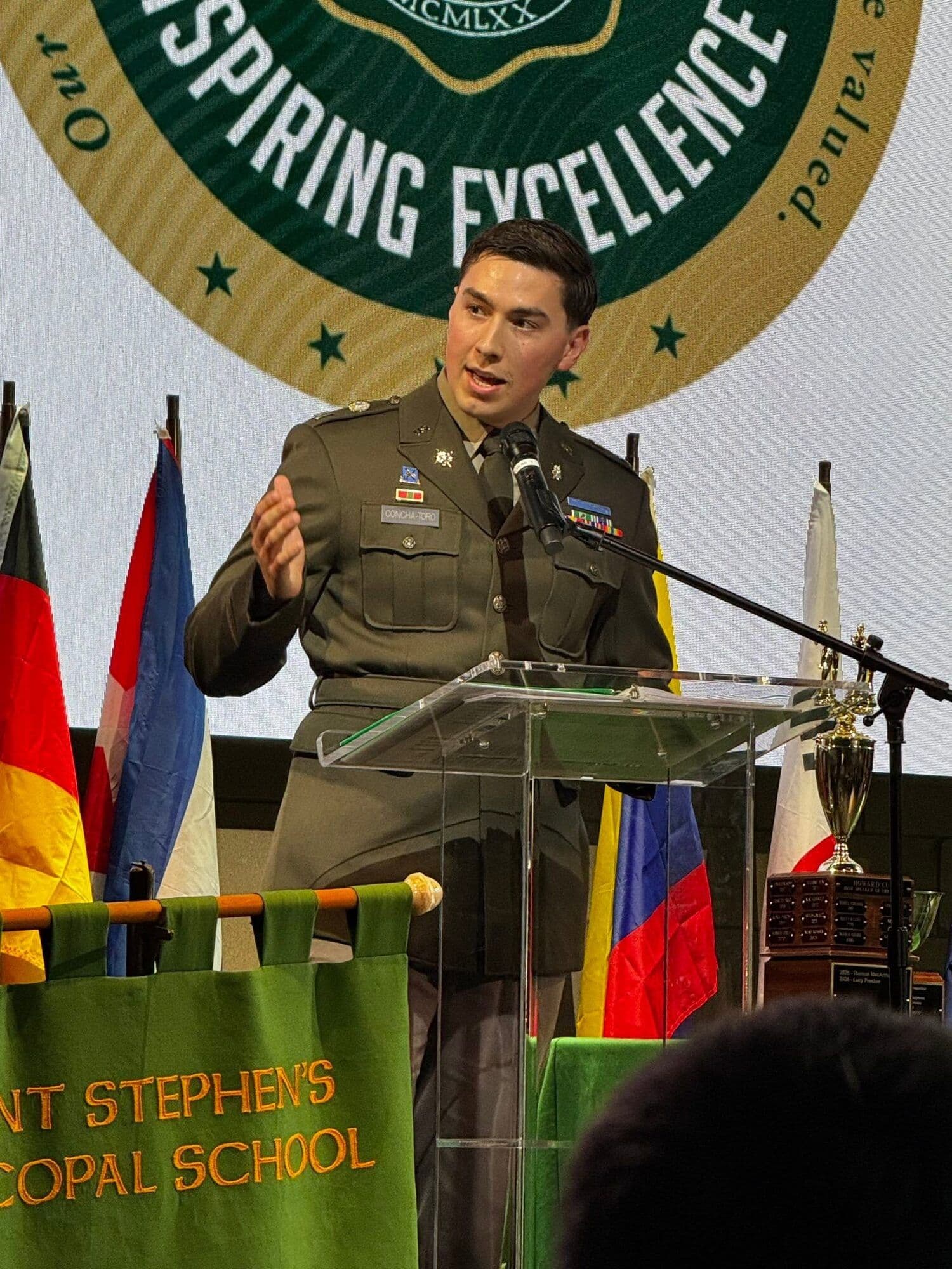 A guest speaker in military dress uniform addresses students from a podium with the Saint Stephen's crest and international flags behind him.